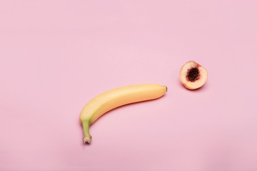 a single yellow banana and a half of a peach representing sexual organs